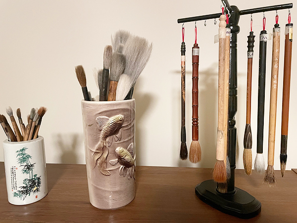 Diana’s stock of brushes. At right, eight brushes hang hair-end-down from the crosspieces of a wooden stand. To the left, a small cup holds smaller brushes and a larger cup holds larger brushes standing hair-end-up.