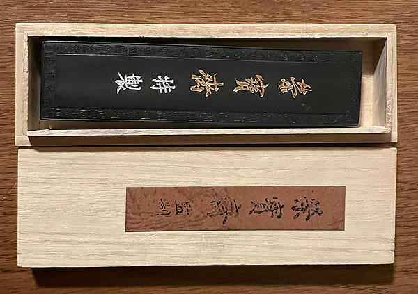 A narrow wooden box holds an ink stick, which appears to be about six inches long by one inch wide, and maybe half an inch thick. The ink stick is black and is embossed with a frame and five gilt Chinese characters.