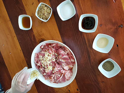The white bowl of pork strips is surrounded by six smaller white bowls holding various ingredients: fish sauce, lemongrass, sugar, oyster sauce, onion powder and black pepper. Ho-Thanh is sprinkling on minced garlic from a seventh small bowl. The tabletop is quite striking, made of alternating planks of light- and dark-colored wood.