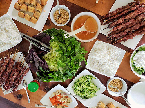 All the ingredients for serving the Grilled Pork recipe are laid out in white dishes on the tabletop. There’s a large bowl of greens in the center (purple basil, mint, cilantro, shredded lettuce), surrounded by two plates of pork skewers, two bowls of rice noodles, two plates of sliced egg rolls, strips of cucumber, pickled daikon (cauliflower) and carrots, chopped peanuts, and a bowl of amber liquid with what looks like peanuts.