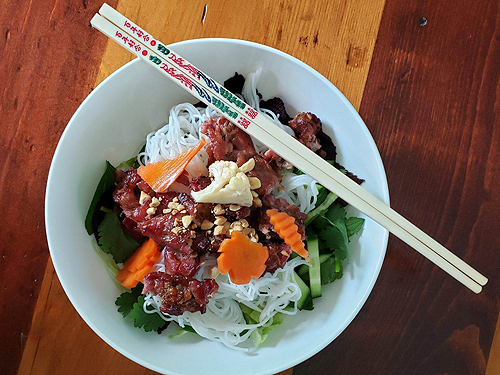 The final presentation of Grilled Pork. A salad bowl contains a layer of greens, a layer of noodles, a heap of pork removed from its skewers, and a sprinkling of peanuts and pickled vegetables. A pair of chopsticks is laid at an angle across the top right quarter of the bowl. Lovely presentation!