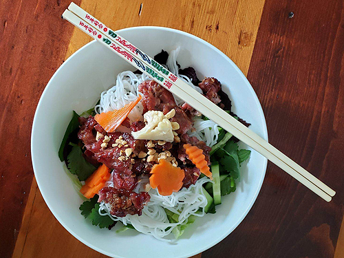 The final presentation of Grilled Pork.  A salad bowl contains a layer of greens, a layer of noodles, a heap of pork removed from its skewers, and a sprinkling of peanuts and pickled vegetables.  A pair of chopsticks is laid at an angle across the top right quarter of the bowl. Lovely presentation!