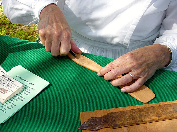 Closeup of Judy’s hands as she starts to carve a design in a spatula handle. Her knife follows a pencil line draft on the wood. She is wearing a white long-sleeved dress in the Mennonite style.