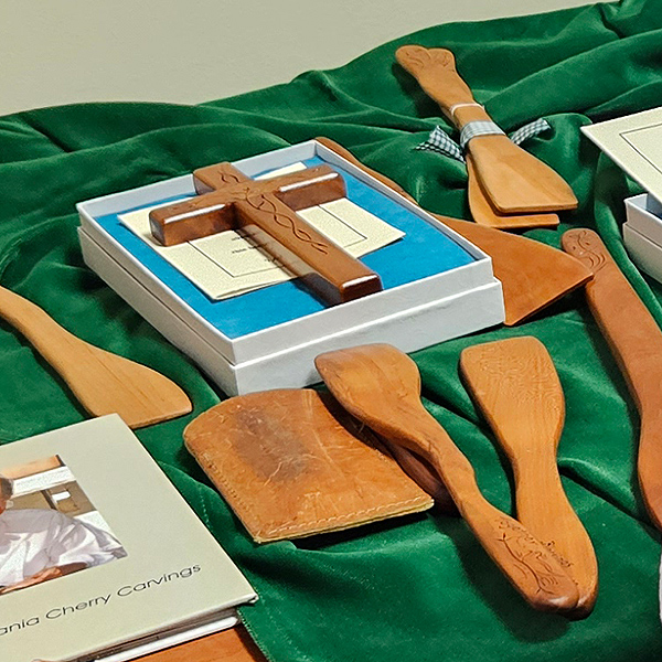 A display of items that the Waltons have made out of cherry wood, decorative folk-art carvings. Laid out on a green velvet tablecloth are several stirring spatulas and a cross in a small box.
