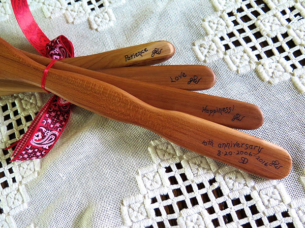 The same four spatulas as above, but flipped over to show the other side of the handle. Each one has an inscription: Patience, Love, Happiness, and 10th Anniversary. They are laid on a handmade lace table runner.