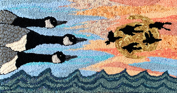 This hooked rug, which appears to be about 3 feet high by 5 feet wide, shows Canada geese in flight over water. At left are three geese: their shoulders are just entering the frame and their heads are about a third of the way across the rug, against sky done in various shades of blue. These geese are the focal point and they dominate the left half of the design. Centered in the right half are five much smaller geese, also flying from left to right, all black, silhouetted against a golden sun and wispy clouds in shades of peach. At the bottom are rows of scalloped waves in shades of dark teal and greenish-gray.