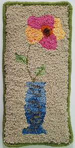 This wall hanging appears to be about 9 inches wide and 18 inches tall, and features a tall blue vase with a single large, colorful flower on a long stem. The design is more primitive than realistic. The flower has five large petals; three are done in shades of magenta, while two others (left and right) are done in shades of gold and peach. The flower center is red, with dark stamens in the very center that are taller than the rest of the loops. The vase is also shaded, dark blue at bottom and fading as it goes up, with an abstract design of darker curlicues decorating it. The background is solid tan. Overhand stitches of dark green yarn bind the edge.