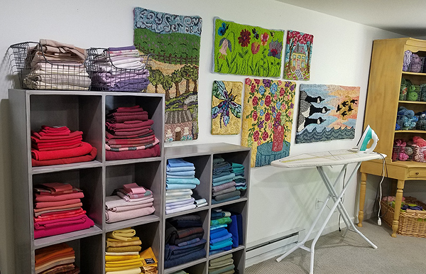 The left half of this image is dominated by gray cube shelves holding neatly folded wool fabrics sorted by color: red, maroon, pinks, blues, browns and golds. There appear to be 14 cubes, with more fabrics in wire baskets on top.  To the right of that stands an ironing board and iron, and behind that, several of Julie’s finished works hang on the wall. At the far right stands a tall maple corner cupboard, holding bundles of colored wool yarn on open shelves, with a large wicker basket underneath.