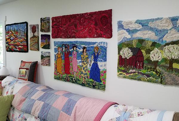 On a white wall in Julie’s studio hang several hooked pieces of various sizes. At the right is Julie’s landscape of Mr. Henry’s barn (described elsewhere in this article). To the left of that, at the top of the frame, is Julie’s abstract work called Red Creek (ditto). Below Red Creek is a piece by Deanne Fitzpatrick, called Coastal Girls, showing five women in colorful dresses against a seashore background. The hangings at far left are too small and far away to be interpreted but seem to be landscapes. The far left piece is another by Deanne Fitzpatrick, called Grey Barn, but that design is dominated by patches of colors representing a hillside. The sofa below these pieces is draped with pastel quilts.