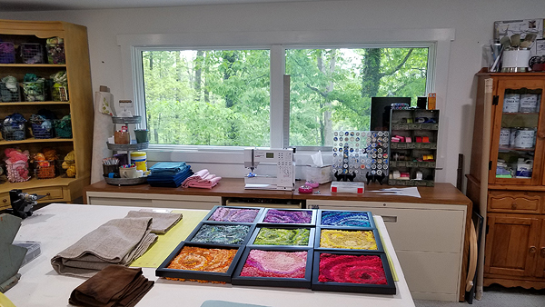 In the foreground is a worktable holding some folded pieces of wool fabric in earth tones, and a hooked piece that is nine squares within black wooden frames. Each of those squares is a different bright color, with lighter and darker tones in abstract patterns. Behind the work table are various cabinets: tall hutches on either side of large windows and wood-topped wide filing cabinets under the windows. The windows look out on Julie’s wooded yard. Colorful rug-making supplies are neatly displayed everywhere.