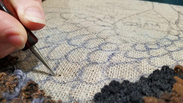 Closeup of Julie making a hooked rug. Most of the frame shows the coarse linen backing material, beige with a pattern of overlapping ovals (short feathers) drawn in marker. The bottom left and right corners are completed areas, with short loops of wool in shades of brown, blue and black. Julie’s right-hand finger and thumb are in the upper right corner, holding the hook down against the fabric. A dark spot just beside the hook is really a widened gap between the threads of the weave; she has just demonstrated how to slip the hook through the fabric.