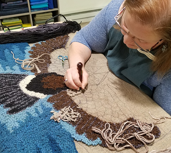 Julie works on her unfinished ‘Goose Moon Rising’ rug, sitting at a frame that holds the backing material taut.  The side further from her is finished, showing the goose’s tail and the sky background.  The side closest to her is empty backing material with outlines sketched on it. She is working in the middle area, filling in the upper part of the goose’s wing. Her left hand is under the rug; her right hand holds a hook as she pulls the fabric worm up through the backing.