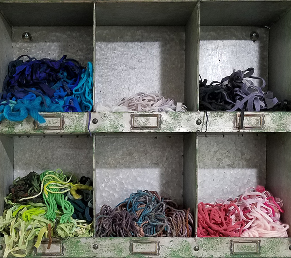 A grid of galvanized tin storage bins hangs on Julie’s studio wall. Each bin is about 8 inches wide and at least that deep, and each holds a heap of skinny wool strips. They’re organized by color. The upper left bin holds blues: navy, teal, sky blue. The bin below that holds greens ranging from dark to bright to pale. Other bins contain pinks, purples, browns, etc. This photo shows six of the nine bins.