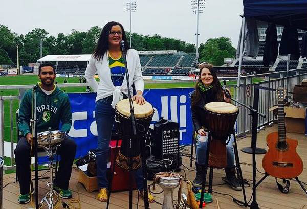 Three musicians ready to perform, surrounded by instruments. Mônica Teles is at center, with her hand on her drum, smiling and ready to speak. Flanking Mônica are a Black man seated behind a snare drum, and a White woman seated behind another Brazilian drum and beside a classical guitar.