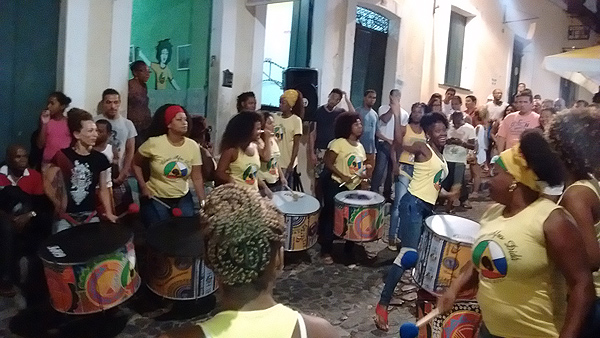 An alleyway in Salvador, Brazil, crowded with onlookers and several female percussionists with large colorful cylindrical drums. The drummers are in a loose circle and a tall young woman is grinning and dancing in the open middle. The women are a range of ages and sizes; all are wearing yellow T-shirts with the school logo. Nearly all of the onlookers and musicians are people of color. It appears to be night-time but the area is lit from above.