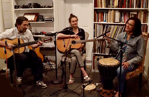 Mônica’s band Suddenly Bossa performs at a house concert.  They are seated close together in front of bookshelves.  At right, Mônica Teles sits with a Brazilian drum between her knees but appears to be playing a shaker instead.  At left, John Catalano plays a classical guitar and smiles.  At center, Jen Catalano holds a guitar but isn’t playing.