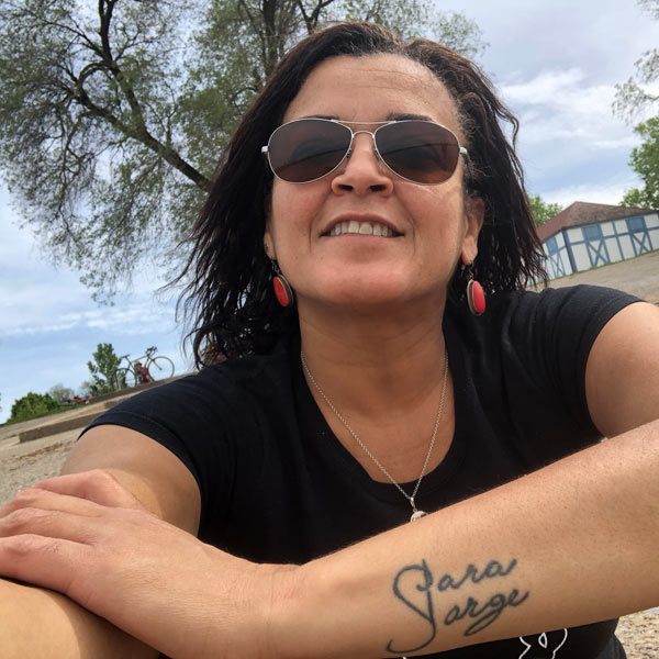 Mônica Teles, in a head-and-shoulders photo, is sitting on pavement with her right arm extended out and her left arm bent in front of her, hand on right elbow, to show off a simple black tatoo on her forearm with the words ‘Sara’ and ‘Jorge’ in script.  She is wearing dark glasses, smiling broadly, and gazing up above the photographer’s head. She’s wearing a black tee, silver necklace, and red-and-brass earrings, and she has dark hair swept back from her face.  The camera is at a 30 degree angle so the horizon is tilted and runs precisely along the top of her shoulders.