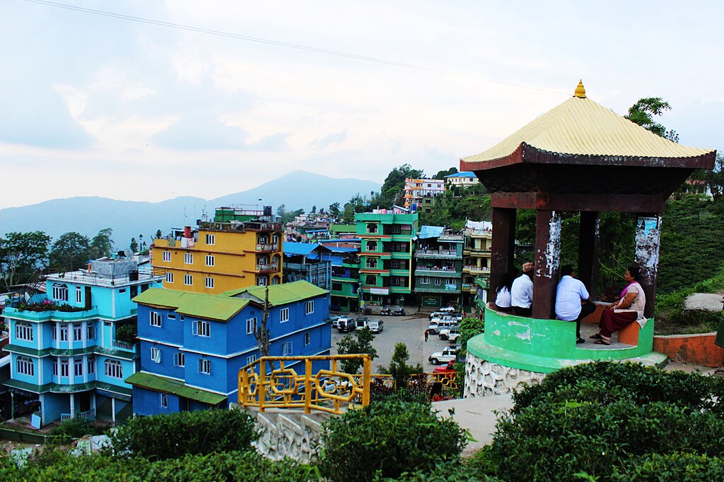 A group of people sit in a small pagoda on the side of a steep mountain. They are at the far right of the photo. To the left, and further down the mountainside, are multi-story houses, small apartment buildings and parked cars. Beyond the houses are tall trees and a view of distant mountains.