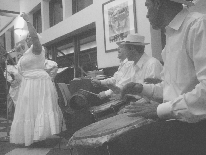 This image from Wikimedia shows a performance of a bomba band in what appears to be a building lobby. There are three drummers to the right, all wearing white dress shirts and white fedora hats. At the left side of the image is a large woman in a long, frilly white dress, gesturing upward with her left arm raised, and grinning.