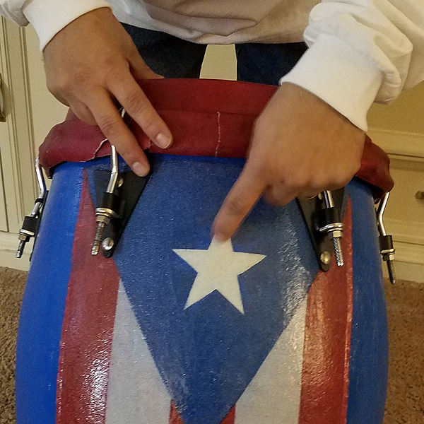 Pedro shows a drum that he made. The body is painted with a Puerto Rican flag; he is pointing to a white star within a blue triangle. Below the triangle are alternating red and white vertical stripes. The drum is about 16 inches in diameter and 3 feet tall, and has a red leather drum-head attached with metal brackets.
