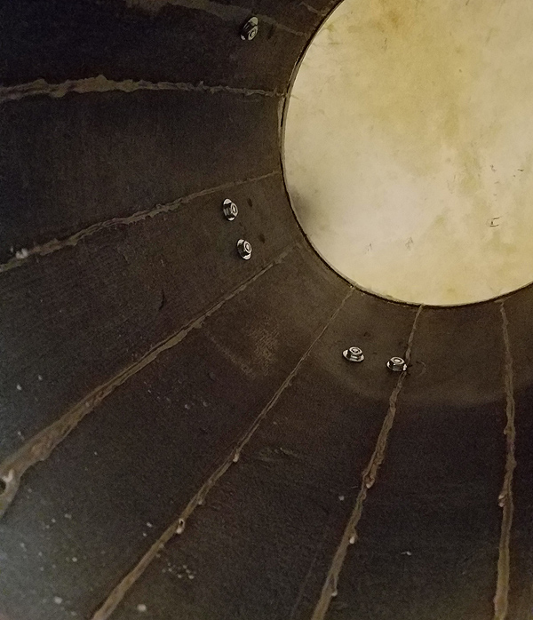 Looking up into a bomba drum from below. The head of the drum appears as a round white circle at the top right of the image, and the barrel-like staves of the drum body are dark wood with lighter-colored sealant, which makes them look like sun rays. A few inches down from the head are bolts which secure the drum-head tuners to the outside.