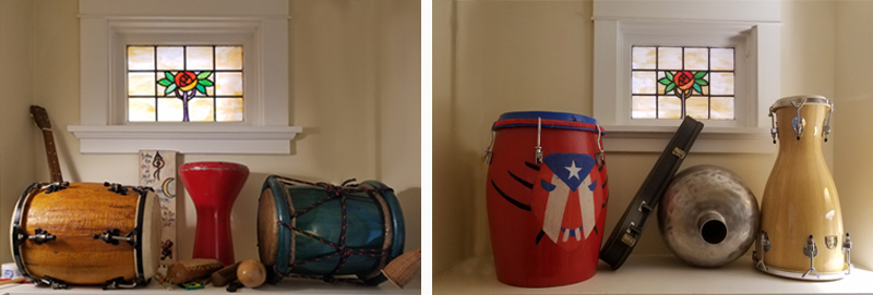 A collection of instruments sits on two deep shelves, with small stained-glass windows above them. There are five large drums, a couple of small guitar-shaped instruments, and some smaller maracas and other percussion instruments.