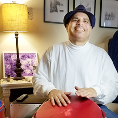 Two Hispanic men with dark skin pose with large Puerto Rican drums in front of a Puerto Rican flag. They wear white shirts and trousers, and white hats with black bands. They are also holding other percussion instruments, including maracas and a basketball-sized shaker covered with colorful fishnet.
