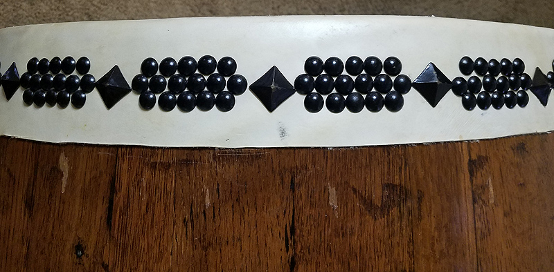 Closeup of the rim of a Taiko drum made by Pedro. The body is dark wood; the head is white leather. Around the rim are black decorative nails in an attractive pattern. Square pyramid nail heads (turned 45 degrees) alternate with arrays of domed-head nails. The square nails are perhaps 1cm on a side. The arrays of domed nail heads (each perhaps 5mm in diameter) consist of a row of six with rows of five above and below; the nail heads touch one another.