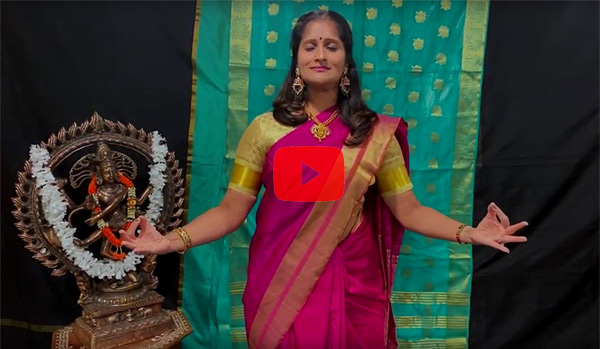 Rachita Nambiar in a still shot from the linked demonstration video.  She stands with her eyes closed; her expression is happy and serene.  Her elbows are by her sides and her hands are held in front of her at rib height, as if she’s holding an imaginary piece of string taut.  Each hand is held with thumb and index finger touching. She looks as though she’s meditating in the lotus pose. She’s wearing a magenta sari over a gold shirt, and she stands in front of a silk drape of aqua silk printed with gilt.  To her right is a piece of Hindu statuary.