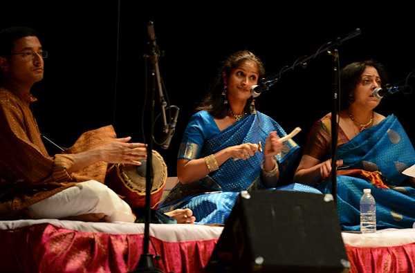Three people sit together on a platform, providing accompaniment for an unseen dancer. The area is dark but the musicians are illuminated. At left, a young man beats a hand drum. At center, Rachita plays tiny hand cymbals; she is wearing a royal-blue sari and wide gold bracelet. At right, a woman older than Rachita, also wearing a royal blue sari, sings into her mic.