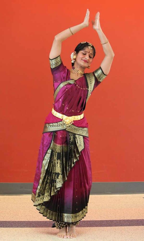 A head-to-toes shot of Rachita performing a dance. She is a slim, pretty woman with dusky skin and dark hair under a black covering, wearing a spectacular silk dress of dark magenta, black and gilt. She is smiling serenely and her eyes are closed. She is standing with her feet together, bending slightly to her left in the dance, arms raised gracefully over her head with the palms a few inches apart.  Her dress is short-sleeved and floor-length, with wide black-and-gilt patterned bands around the arms and arcing up the sides of the bodice, as well as on the skirt. Sharp pleats make a triangle from her waist to her left shoulder. The skirt is a wrap design with at least 25 pleats in each of two layers. The top layer arcs up from floor to hip in many, many pleats, repeating the black-and-gilt pattern. Below knee level, the bottom layer of the skirt fades gradually from purple into black-and-gilt at the hem.