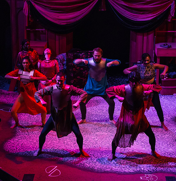 Seven dancers spotlighted on a dark stage. Most of the costumes are tights and long tunics that vary in color and style. Four of the five in front wear chest plates resembling armor. The two dancers in front and three behind them stand with their feet more than a yard apart, knees slightly bent, arms held straight out to the sides at shoulder level and clenched fists out in front of them. Behind those dancers and to the left, two women appear to be seated; one is grinning with her head thrown way back; the other is gazing across the stage behind the dancers.