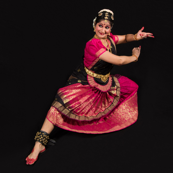 Rachita dancing in a studio shot against a solid black background. Her torso is upright but her knees are bent so she’s close to the ground, with her right leg extended to show a broad band of little bells buckled around her ankle, and her bare foot with red paint on toes and sole.  She’s looking to her right, smiling coyly and cutting her eyes even further right, but her arms are held out to her left, at shoulder level.  Her elbows are bent and her hands are close together, fingers extended in a complex gesture. Her fingertips are painted red. Her dress is bright magenta pleated silk with gilt overprinting and black on the bodice front and behind her hips.  She’s wearing gold bracelets, a gold metal belt, a black hair-covering with gold and white ornaments, and a large maroon bindi low on her forehead.