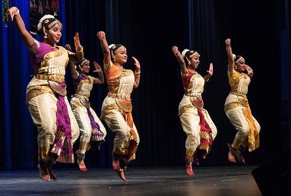 Five teenage girls are dancing onstage, wearing saris of silver and gold silk and ornamented black hair coverings.  Each girl wears a different jewel-toned tee-shirt and matching sash: magenta, purple, orange, red and saffron. They are all jumping up, about a foot off the ground, showing red-painted toes and bell bands around their ankles. Each is holding her right arm up at an angle with wrist bent and fingers together so the hand forms a teardrop shape. Each is holding her left arm with elbow bent, palm near her face and palm facing out, displaying red-painted fingertips and a red dot on the palm.  They are all girls of color, though it’s not clear whether all are of Indian descent.