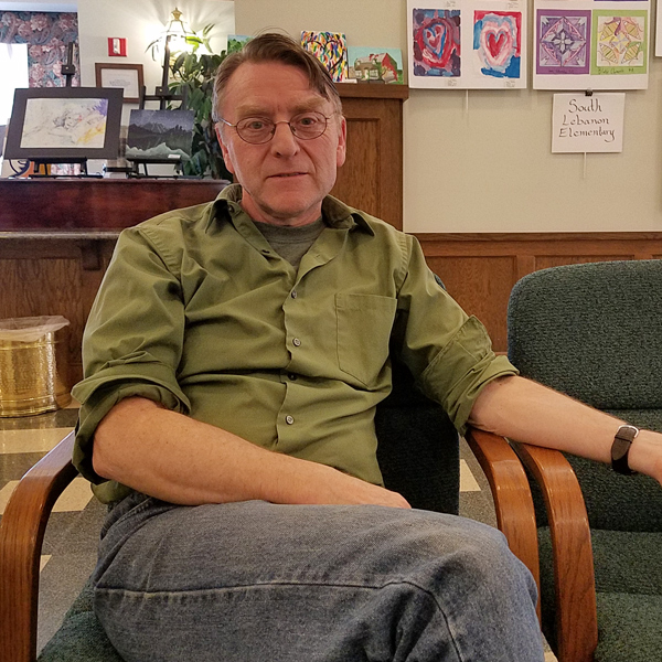 Seamus Carmichael in a candid shot. He’s a white man who appears to be in his 60s, wearing glasses, jeans, and an olive-green shirt.  He looks relaxed and interested.  He appears to be sitting in a lobby.