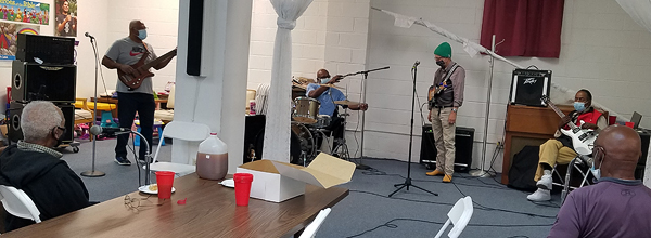An informal rehearsal in the church’s fellowship hall. Rev. Jackson and Mac McFarland are singing from their seats at a table in the foreground; they are sitting with their backs to the camera. On the far side of their large circle, four others are playing instruments: Wayne has his electric bass guitar, Anthony is sitting behind a drum kit; Tom has his mandolin, and Mark has his electric guitar. All the men are wearing masks and staying several feet apart from one another.