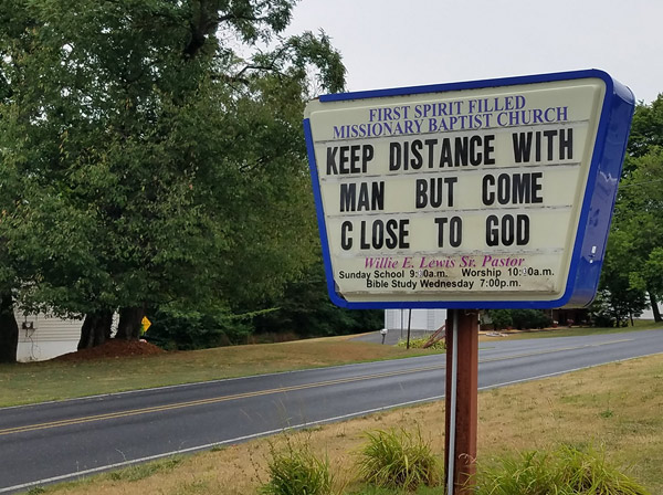 Trapezoidal sign by the road in front of the church: the name of the church appears at top, worship details at the bottom. In the center, movable letters create this message: “Keep distance with Man but come close to God.”