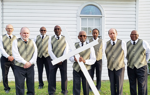 The Spiritual Messengers pose in front of their church. Rev. Moses Jackson holds a large white cross in front of him, at an angle with one end on the ground. The cross is made of 4-by-4 material and is taller than Rev. Jackson.