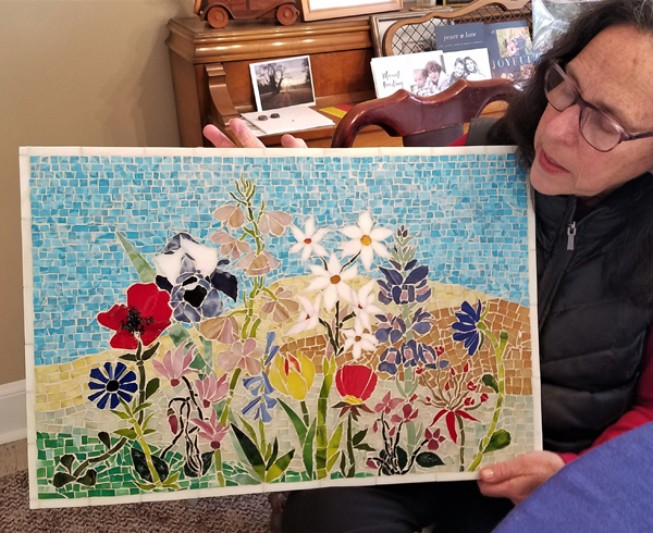 Susan Leviton holds a glass mosaic she made.  It is a rectangle with a couple dozen different flowers all growing together in a garden, against a background that suggests a distant hillside and a patch of sky.