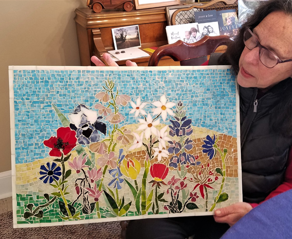 Susan Leviton holds a glass mosaic she made. It is a rectangle with a couple dozen different flowers all growing together in a garden, against a background that suggests a distant hillside and a patch of sky.