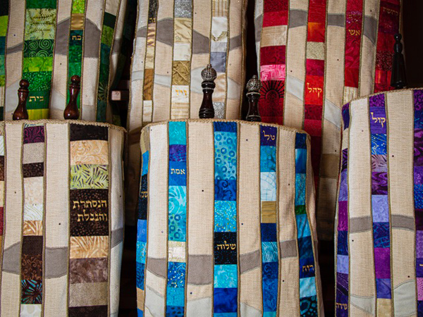 The Torah scrolls themselves appear to be about 30 inches tall, and are stored vertically. The mantles are quilted in stripes: alternating colored strips (some with Hebrew writing) and lighter beige strips. The design echoes the sculptural cabinet that houses the Ark.