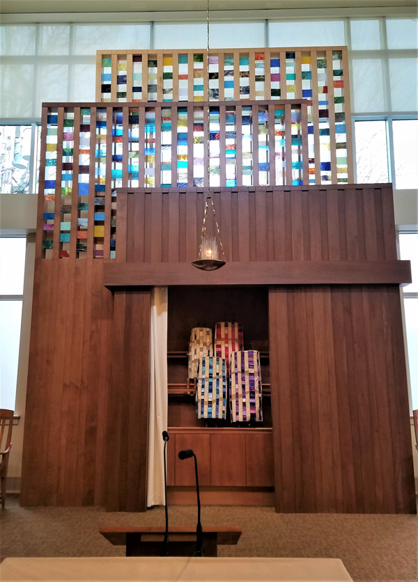 Behind a lectern, a tall sculptural structure houses the congregation’s Torah scrolls, which draped with mantles made by Susan Leviton. The bottom part of the structure is vertical wood paneling; this transitions into vertical strips of stained glass separated by wood laths.