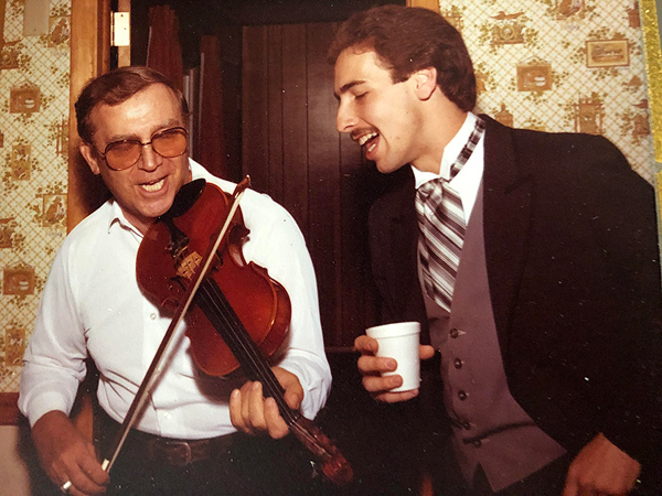 George Caba is a middle-aged man, somewhat stout, wearing a white buttoned shirt and large aviator-style glasses. He is playing fiddle and singing. Lenny Tepsich is at right, singing with his eyes closed. He’s wearing a black tuxedo jacket, white wing-collar shirt, a brown vest, and a brown-and-white striped necktie that’s much wider than a standard tie. The feel of the photo suggests the 1970s.
