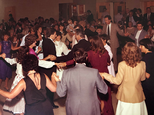 Kolo dancers pack a large room at a church, smiling and looking like they’re having fun. They are all wearing dressy clothes (they are at a wedding reception) and holding hands in lines or circles. Clothes and hairstyles suggest the 1970s.