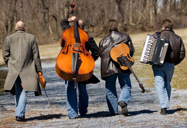 The four members of Sviraj walking away from the camera, on a flat gravel road, with their instruments on their backs. They are wearing winter coats, and the vegatation in the background is leafless.