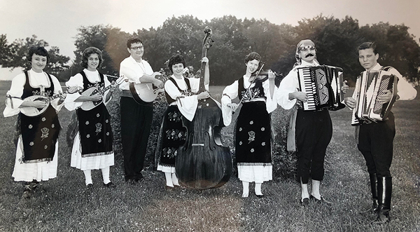 Black-and-white photo of a tambura band, all wearing traditional Serbo-Croatian clothing. The four women wear dark embroidered pinafores over white dresses; the three men wear white shirts and black pants. From left to right, there are three players of stringed instruments; a woman with upright bass in the middle; a fiddler; a man with an accordion, wearing a head scarf, sash, and pants that show his ankles. On the far right is teenage Danilo with his accordion, pants tucked into shiny knee-length boots.