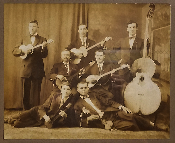 Seven men pose with tamburitza instruments in a sepiatone photo from the early 20th Century. They are all wearing suits and ties; they are all clean-shaven white men who appear to be in their 30s to 50s. Two of them are seated in the center; three are standing behind them; two are on the floor in front, lying on their sides and propped on one elbow, with their heads close together.