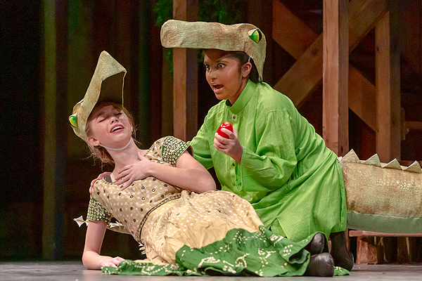 In this still photo from the Panchatantra Tales production, two young girls are wearing alligator costumes.  One is swooning on the floor propped on one elbow, while the other is kneeling beside her, supporting her with an arm, and holding a bright-red fruit. The second girl has a wide-eyed expression of intent. They are clearly having fun acting their parts.