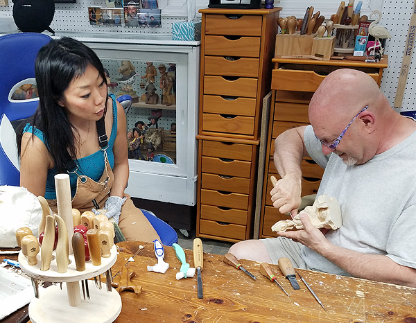 Aron Rook and Jim Hiser sit together behind a work-table. Aron is a young woman of Asian appearance, with straight black hair. Jim is a portly white man who appears to be in his 60s, with a shaved head and a short white goatee. He is working on a wood-carving held in his lap.