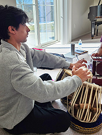 A dark-haired teenage boy in gray hoodie plays tabla drums, using his fingertips and the heels of his hands.  The drums are about 12 inches across and 20 inches high.  They have skin drum-heads and white cords running vertically, with short,thick wooden rods stuck between drum and cords to increase tension on the drum heads.